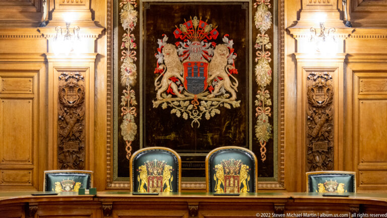 Hamburg coat of arms above high-backed civic chairs framed by carved wood panels and floral textile ornament.