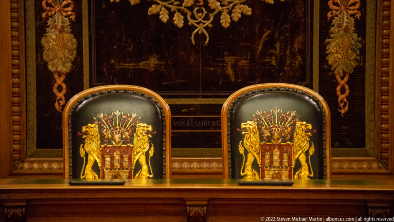 Two high-backed chairs bearing Hamburg’s coat of arms beneath a wall panel with floral ornament and lettering.