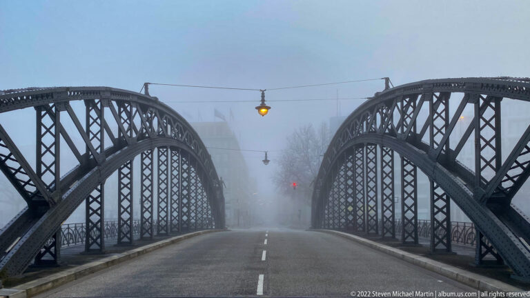 Empty roadway crossing a twin-arched riveted steel bridge, with overhead wires and a hanging lamp fading into fog.
