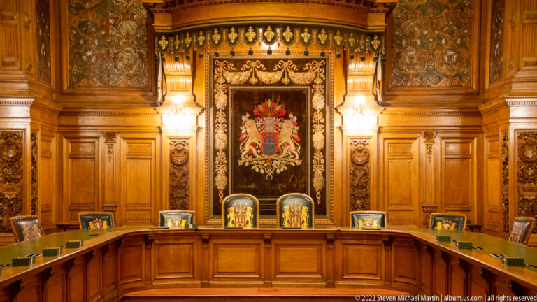 Wide ornate Hamburg Rathaus council room with curved desks, carved woodwork, central coat of arms, and ceremonial chairs.