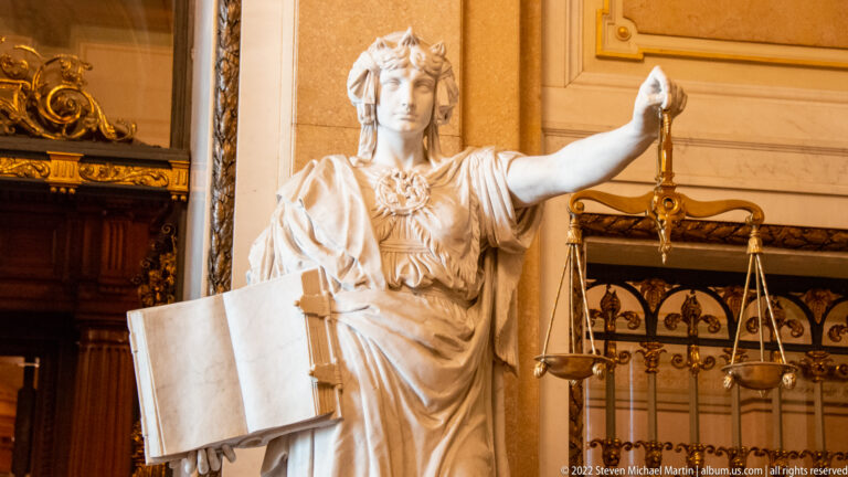 Pale stone female figure of Justice holding balance scales and an open book beside an ornate Rathaus doorway.