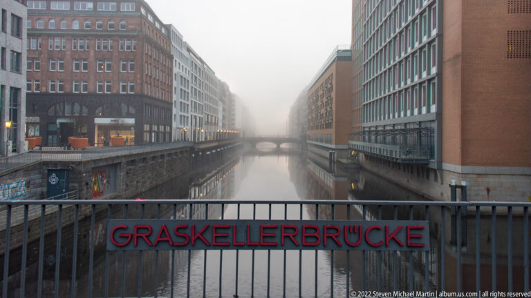 Foggy canal viewed across a railing labeled “GRASKELLERBRÜCKE,” with buildings lining both sides.