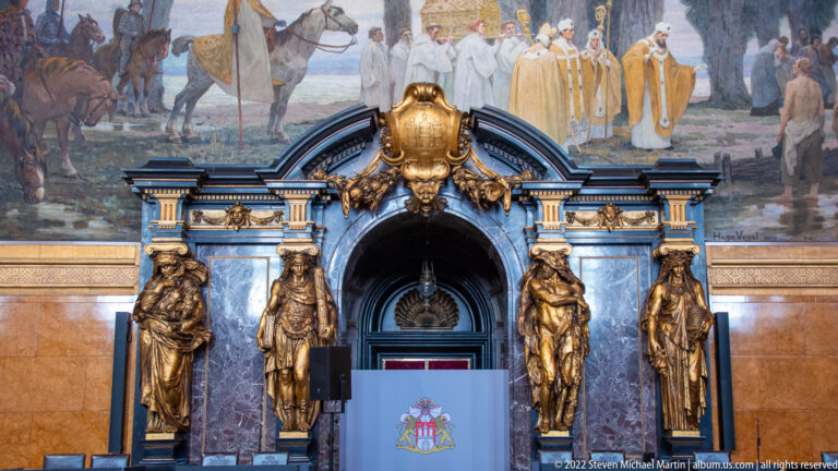 Blue-and-gold arched portal with four sculpted figures beneath a mural of clergy and a ceremonial procession.