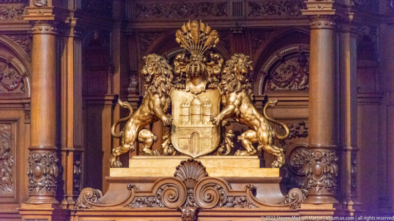 Gilded Hamburg coat of arms with two rampant lions and fan-like crest mounted against carved dark wood.