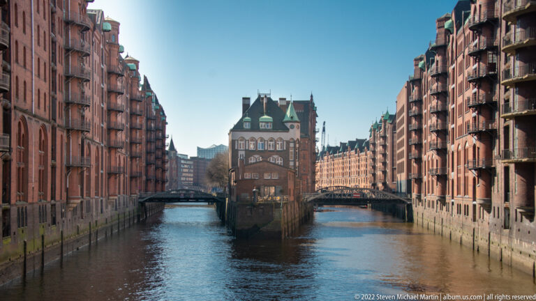Wasserschloss centered between two canals and flanked by red-brick Speicherstadt warehouse blocks under a clear blue sky.