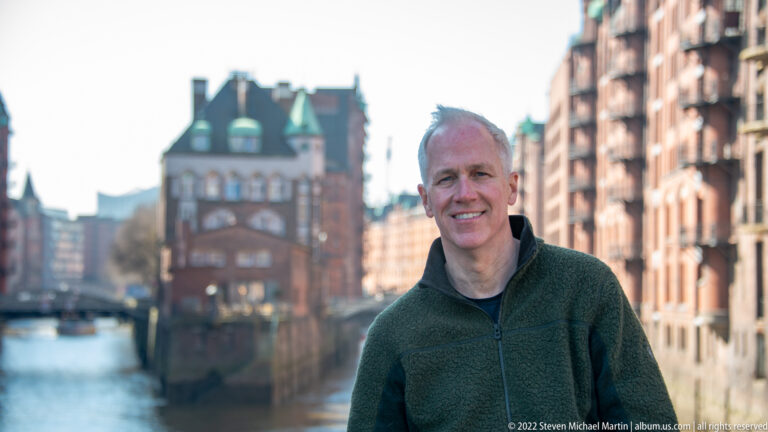 Smiling adult man in foreground with the Wasserschloss and red-brick Speicherstadt warehouses softly blurred behind him.