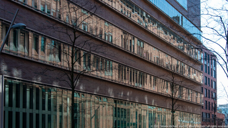 Long modern brick-and-glass building with repetitive horizontal windows, glazed ground floor, and bare trees along the façade.
