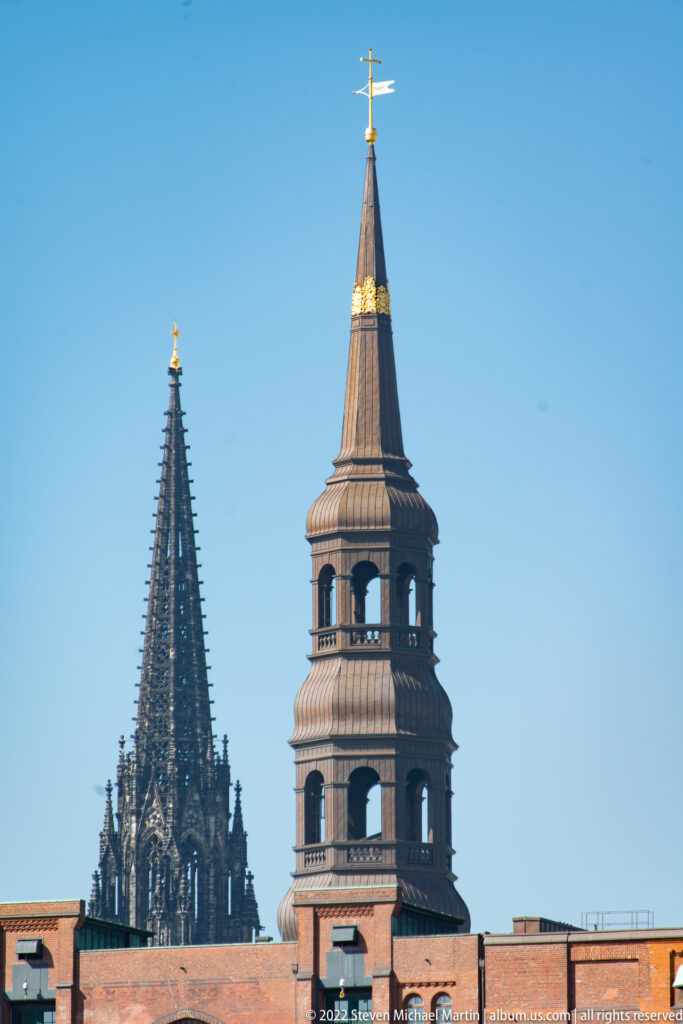 Dark Gothic spire beside a broader open-belfry church tower rising above a low brick roofline.