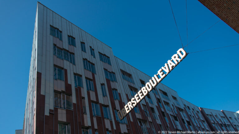 White suspended ÜBERSEEBOULEVARD letters stretch diagonally above modern HafenCity buildings against a deep blue sky.