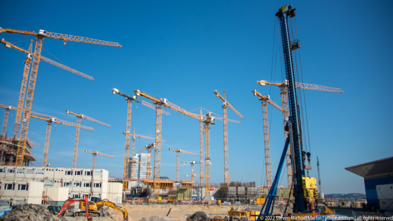 Numerous tower cranes, construction equipment, and unfinished concrete structures fill a large Hamburg redevelopment site beneath blue sky.