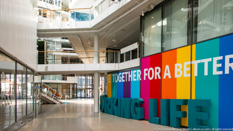 White modern office atrium with glass balconies and a colorful installation reading “TOGETHER FOR A BETTER WORKING LIFE.”