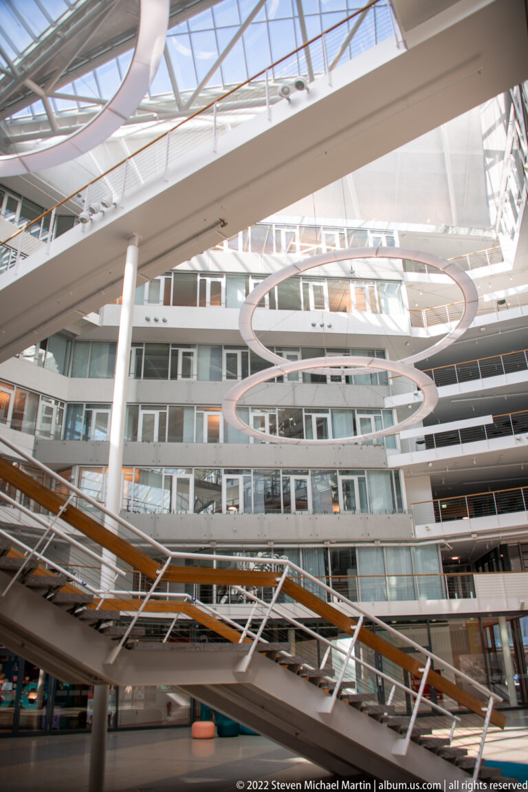 Large glowing circular light rings suspended inside a multi-level atrium with balconies, glass railings, and exposed framing.