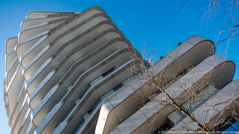 Low-angle view of a modern high-rise with broad stacked white balconies curving around its façade against blue sky.