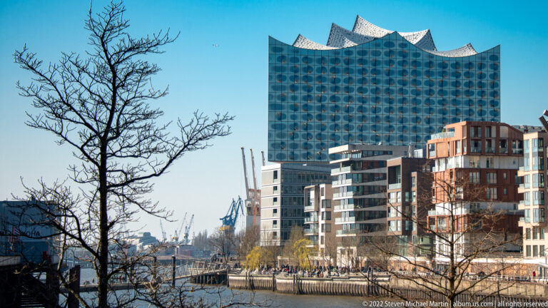Elbphilharmonie rising above modern HafenCity buildings, bare tree branches, waterfront paths, and distant harbor cranes.