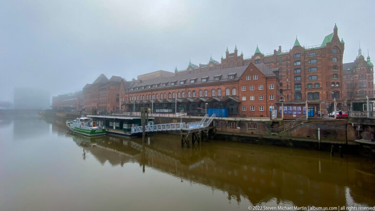 Foggy Speicherstadt canal lined with red-brick warehouses and a green-and-white vessel moored beside the quay.