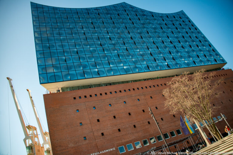Low-angle Elbphilharmonie view with red-brick base, blue glass wave-shaped upper structure, and harbor cranes at left.