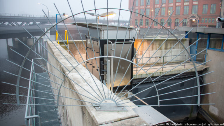 Fan-shaped steel safety grille around a concrete harbor structure, with yellow railings and dock equipment beyond.
