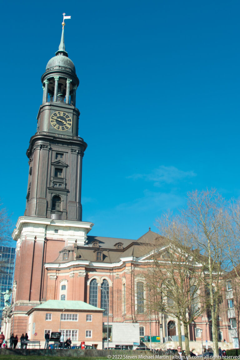 Side view of St. Michael's Church with broad brick nave and tall clock tower rising against blue sky.