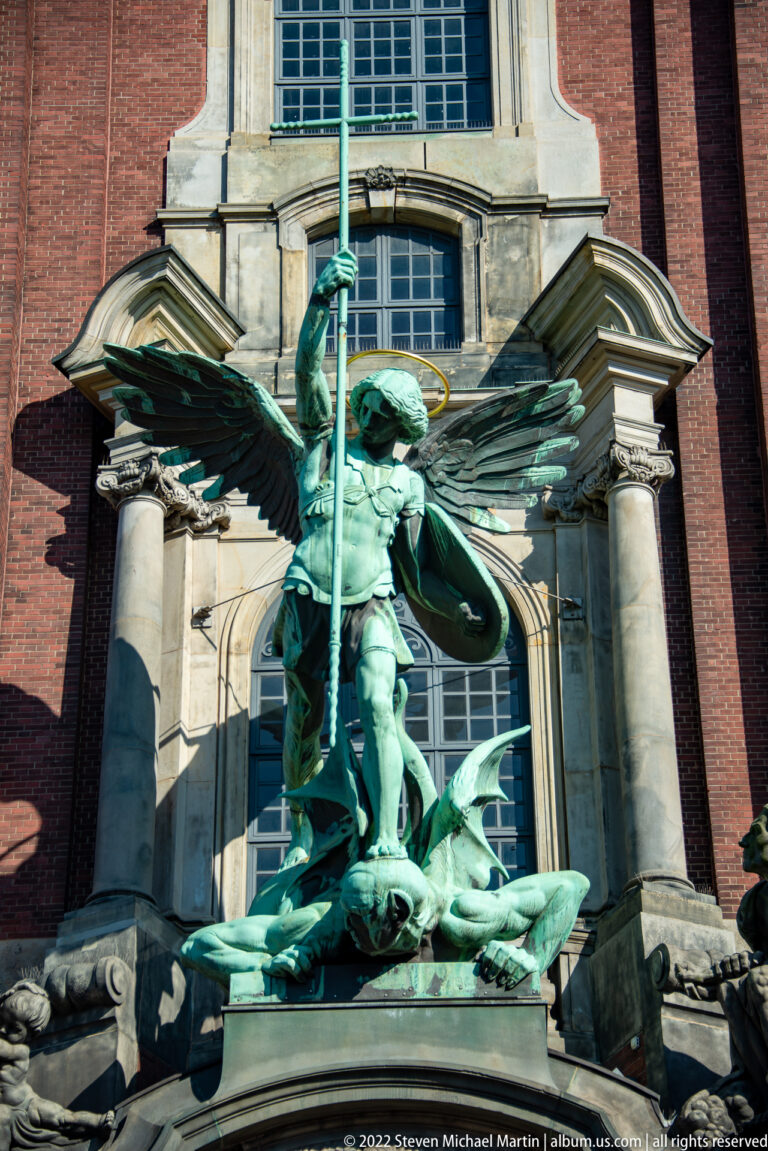 Green bronze Archangel Michael with spread wings and spear defeating a fallen devil above the church entrance.