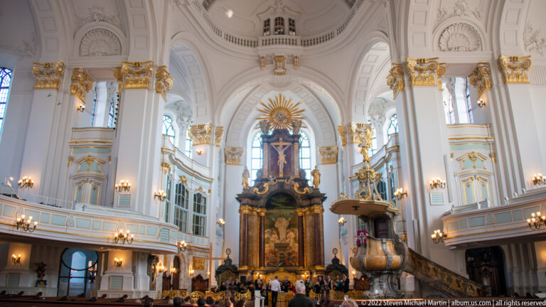 White-and-gold interior of St. Michael's Church with monumental altar, galleries, pulpit, crucifix, and worshippers.