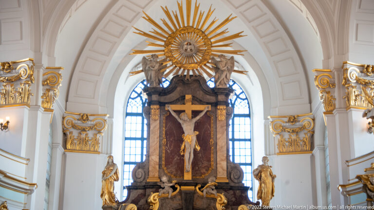 Large crucifix beneath a radiant golden sunburst within the ornate white-and-gold altar of St. Michael's Church.