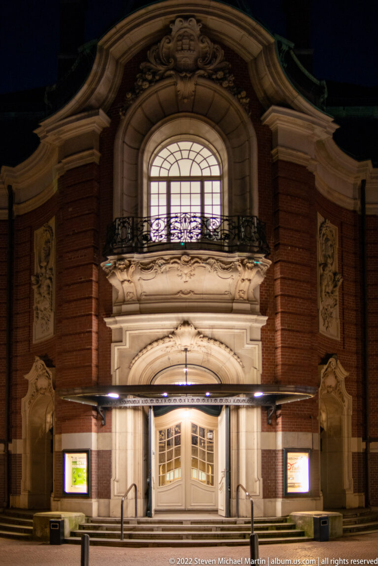 Night view of the Laeiszhalle entrance with large arched window, stone ornament, balcony, and warm lighting.