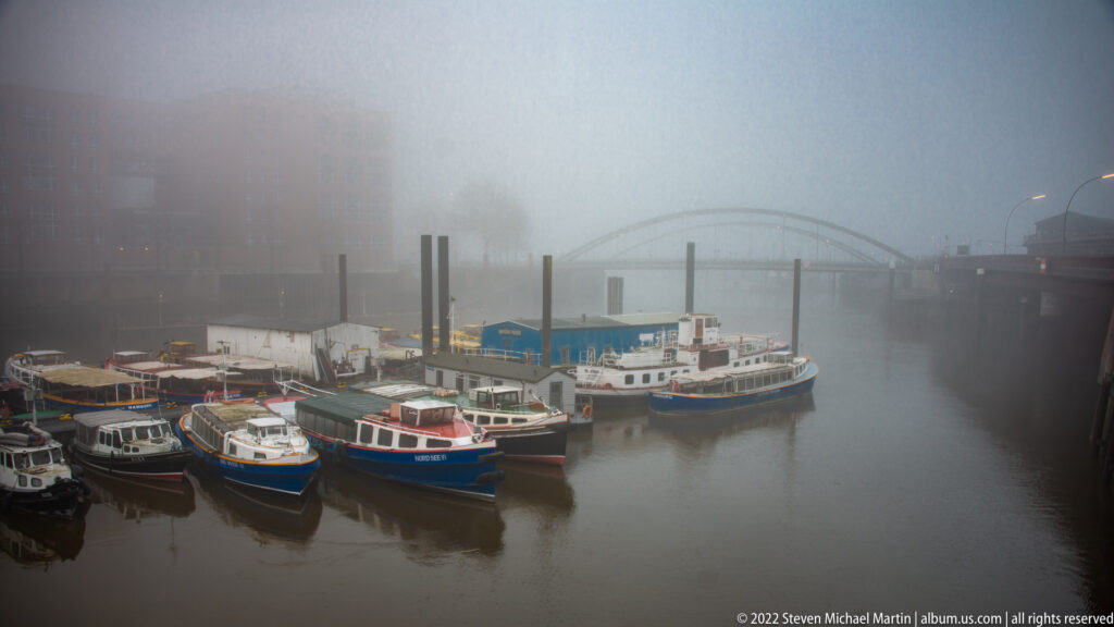 Small boats moored in a foggy Hamburg harbor basin with vertical pilings and a faint bridge in the distance.
