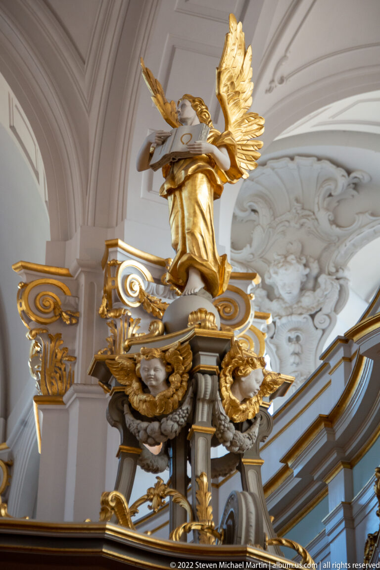 Gilded winged figure above sculpted masks and scrollwork on an ornate white-and-gold church interior element.
