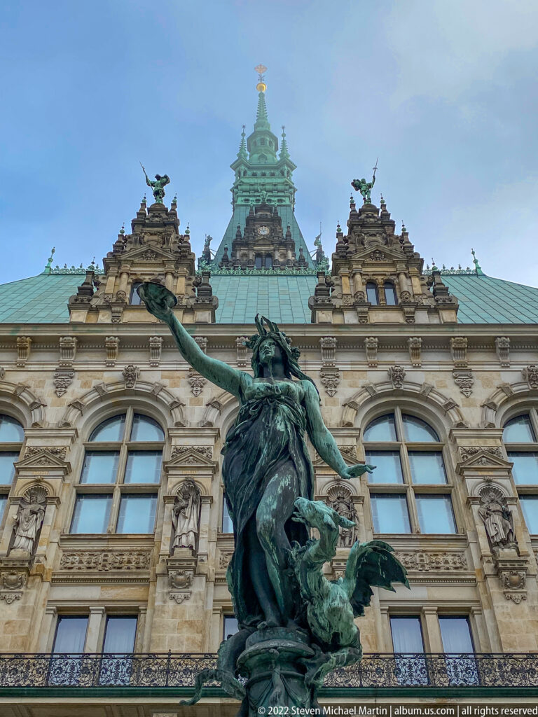 Green bronze Hygieia figure with extended arm and coiling serpent before the ornate Hamburg Rathaus courtyard façade.
