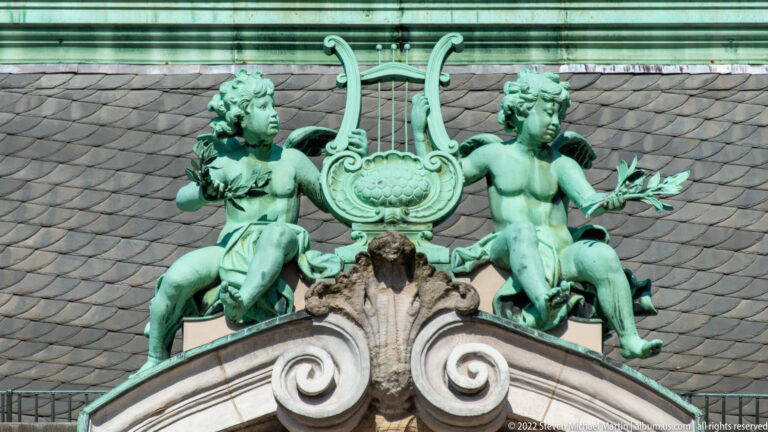 Two green-patinated seated figures flank a large lyre above a stone pediment on Hamburg Rathaus.