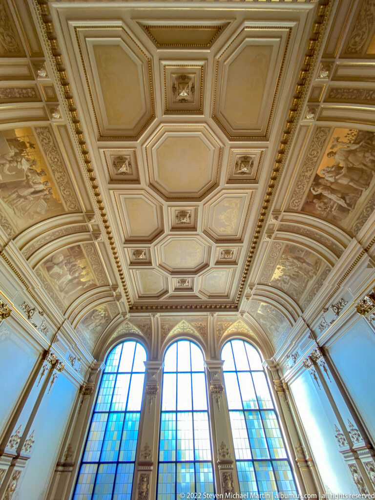 Ornate Hamburg Rathaus ceiling with geometric coffers, painted panels, gilded borders, and tall arched windows below.