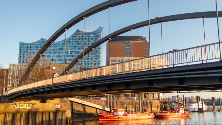 Dark arched bridge over harbor water with red workboats below and the Elbphilharmonie visible in the background.