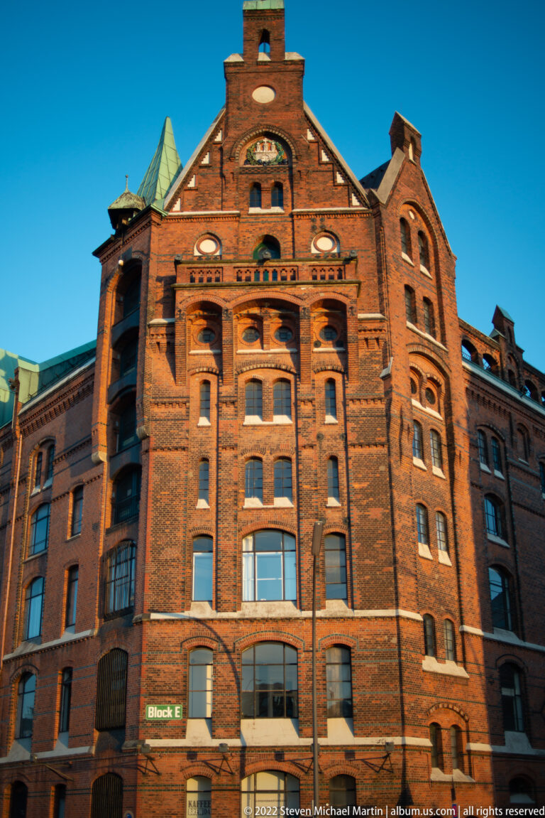 Red-brick Speicherstadt warehouse labeled Block E with stepped gables, arched windows, and decorative brickwork.