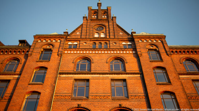Sunlit upper façade of a Speicherstadt warehouse with stepped central gable, arched windows, and decorative brick patterns.