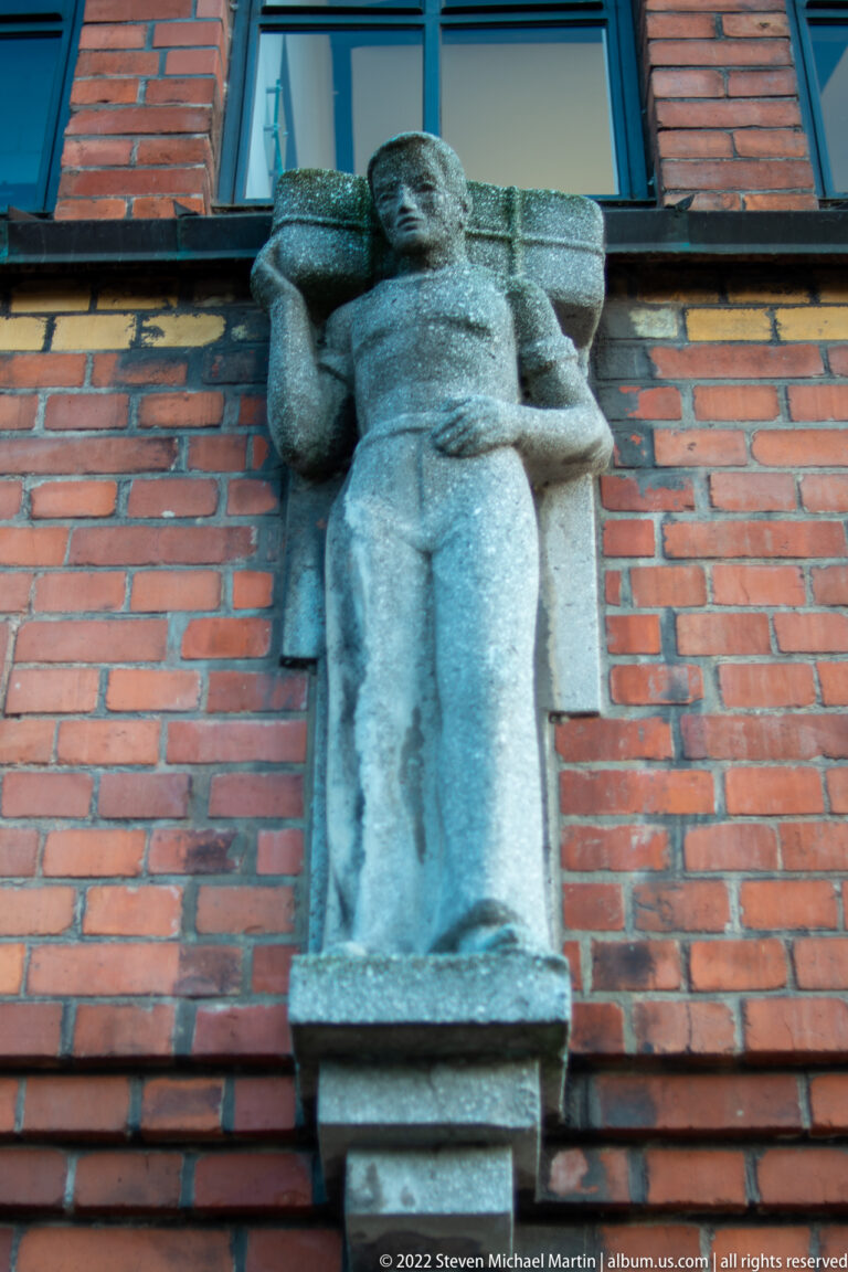 Weathered stone figure carrying a heavy bundle across the shoulders, mounted on a red-brick Speicherstadt warehouse façade.
