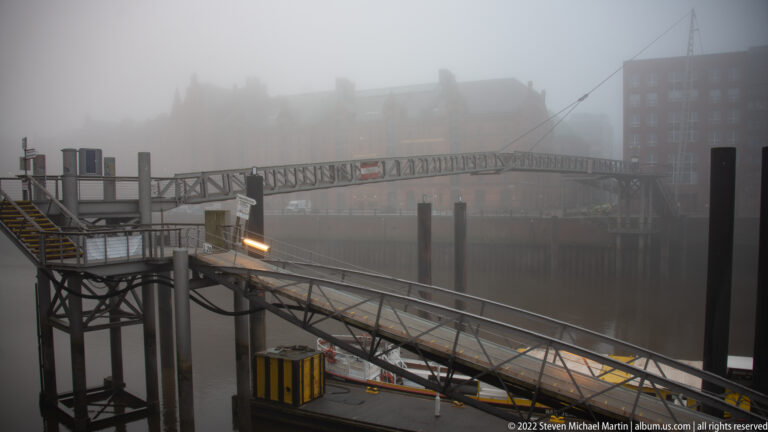Metal harbor gangways, stairs, pilings, and platforms suspended over dark water in heavy fog.