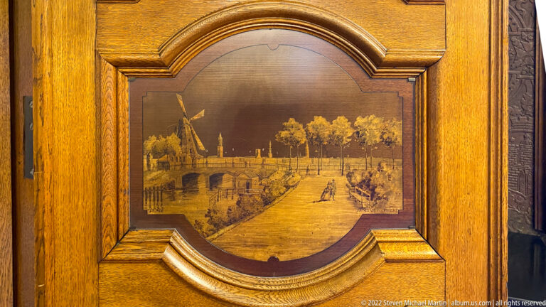 Framed wooden inlay showing a windmill, bridge, canal, trees, and small figures in a historic waterfront scene.