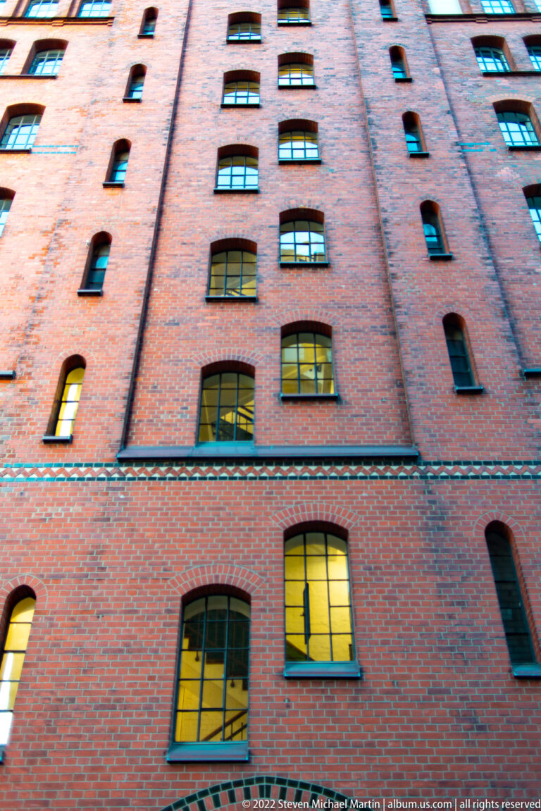 Steep upward view of a tall red-brick Speicherstadt warehouse façade with narrow windows and several glowing lower openings.