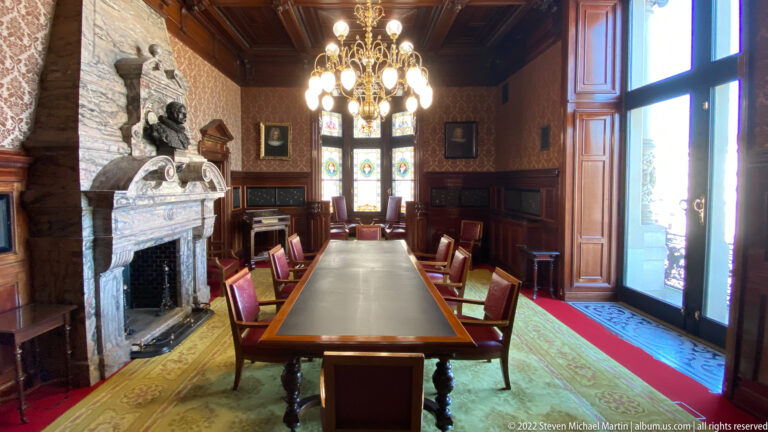 Long meeting table with red chairs beside a carved stone fireplace in a dark wood-paneled Rathaus room.