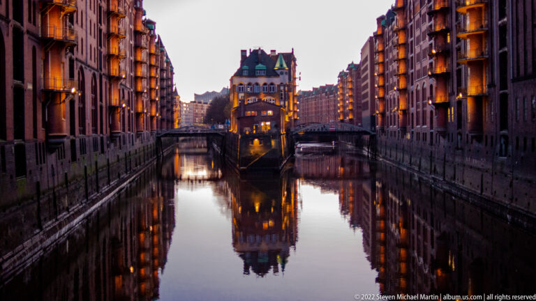 Wasserschloss glowing at dusk between illuminated Speicherstadt warehouses, with buildings reflected in calm canal water.