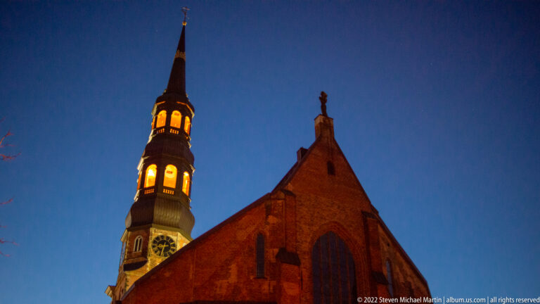 Illuminated brick church tower with clock faces and slender spire beside a steep gabled nave at dusk.