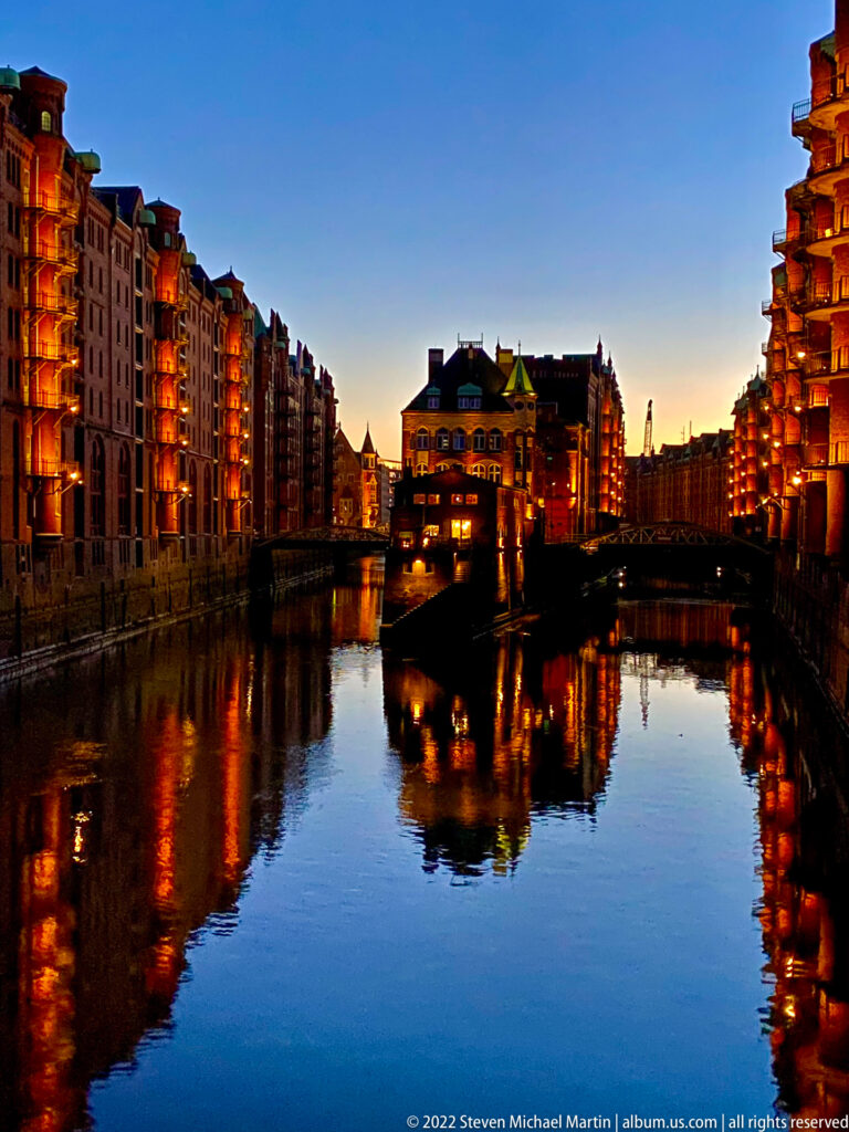 Wide twilight canal view of Wasserschloss surrounded by illuminated Speicherstadt warehouses and warm reflections on the water.