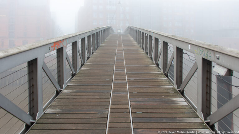 Wood-plank pedestrian bridge with gray steel railings extending straight into dense fog.