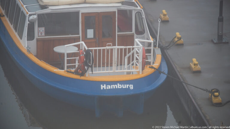Close view of blue-and-yellow harbor vessel bow labeled “Hamburg,” tied beside yellow dock fittings in fog.