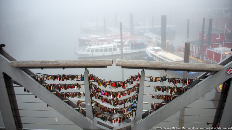 Rows of love locks attached to a harbor railing, with passenger boats and pilings blurred in fog below.