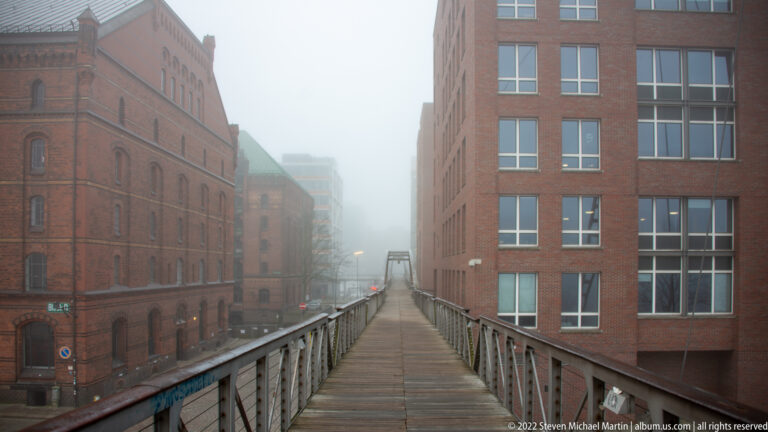 Wood-deck pedestrian bridge between tall red-brick buildings in a foggy warehouse district.