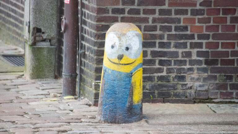 Concrete bollard painted as a yellow-and-blue Minion-style character beside a brick wall and cobbled pavement.