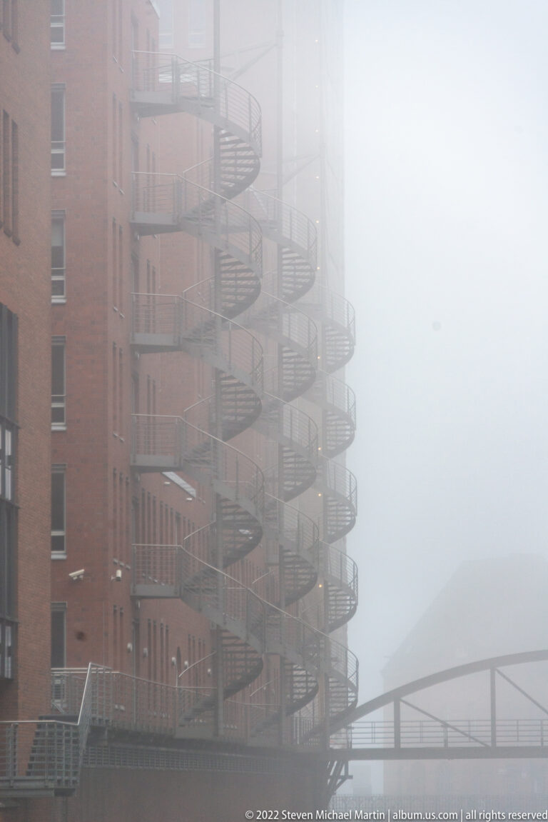 Series of spiral metal exterior staircases climbing a red-brick warehouse and fading into fog.