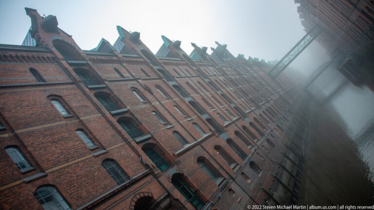 Tilted view along tall gabled red-brick Speicherstadt warehouses beside a canal in fog.