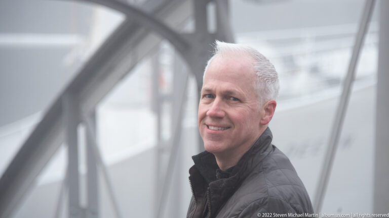 Smiling man in dark jacket beside a metal bridge railing, with foggy waterfront structures blurred behind.